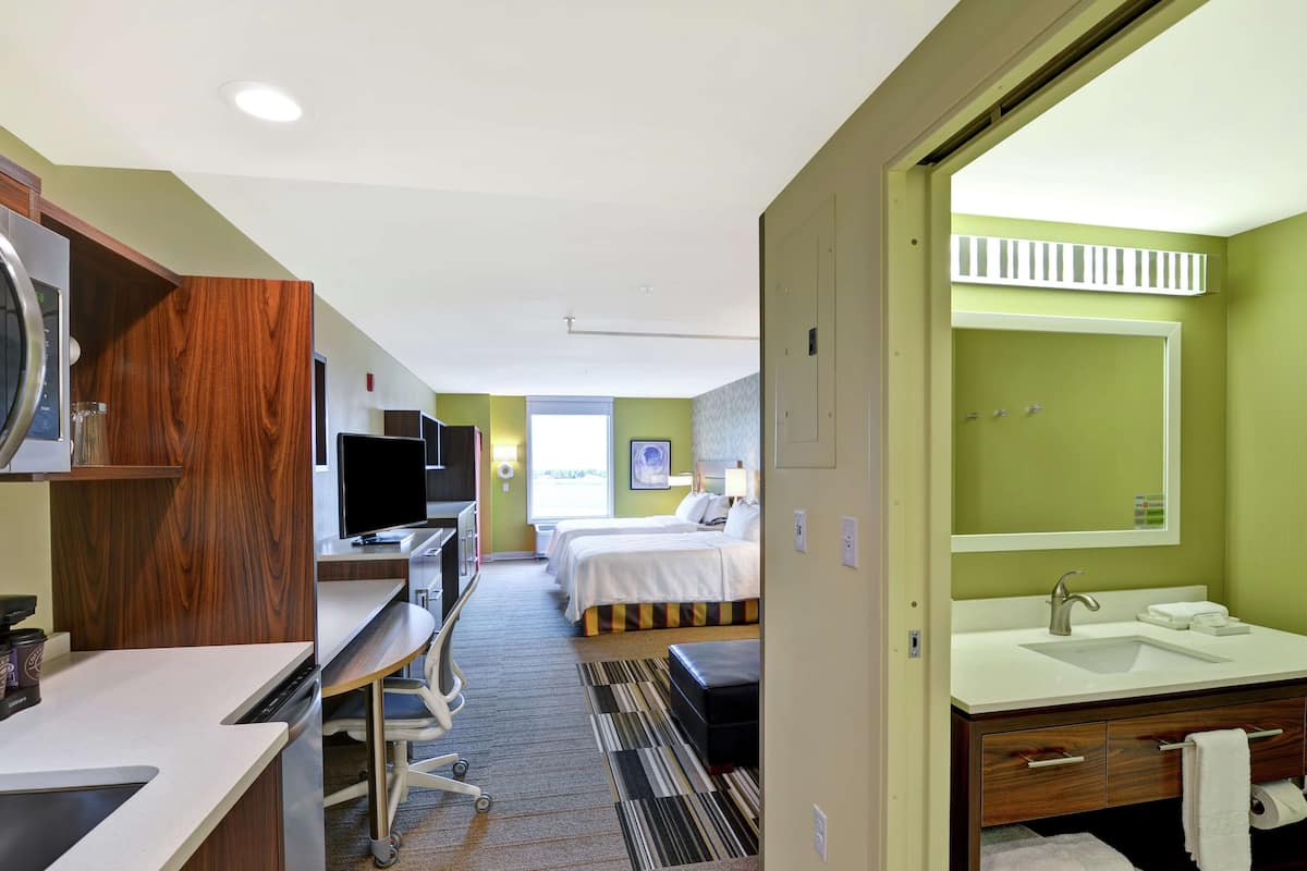 A hotel room with a desk, two beds, a bathroom with a sink and mirror, and a view of the outdoors.