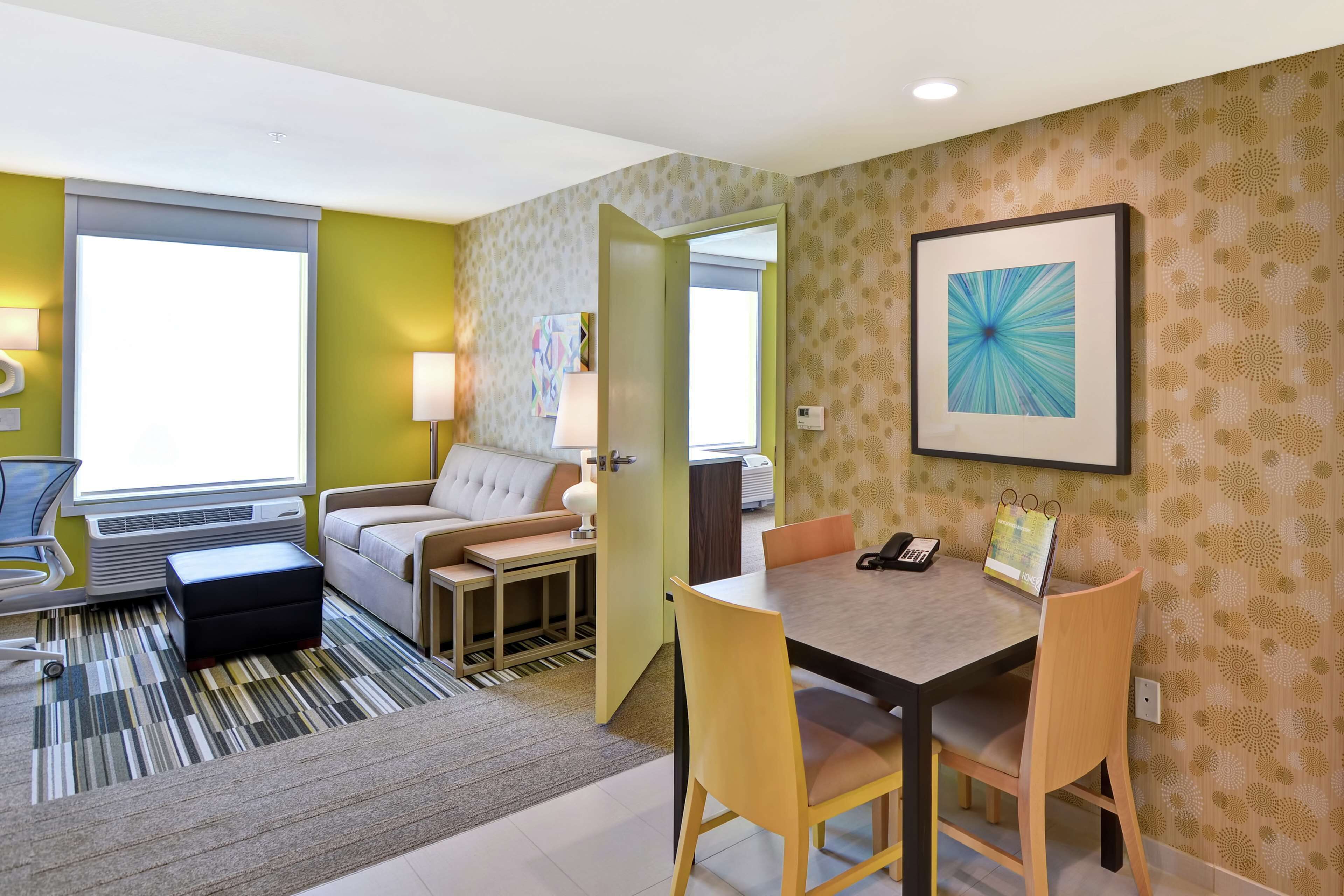 A hotel room with a sofa, a dining table with chairs, a small table with a lamp, a radiator, and a window.