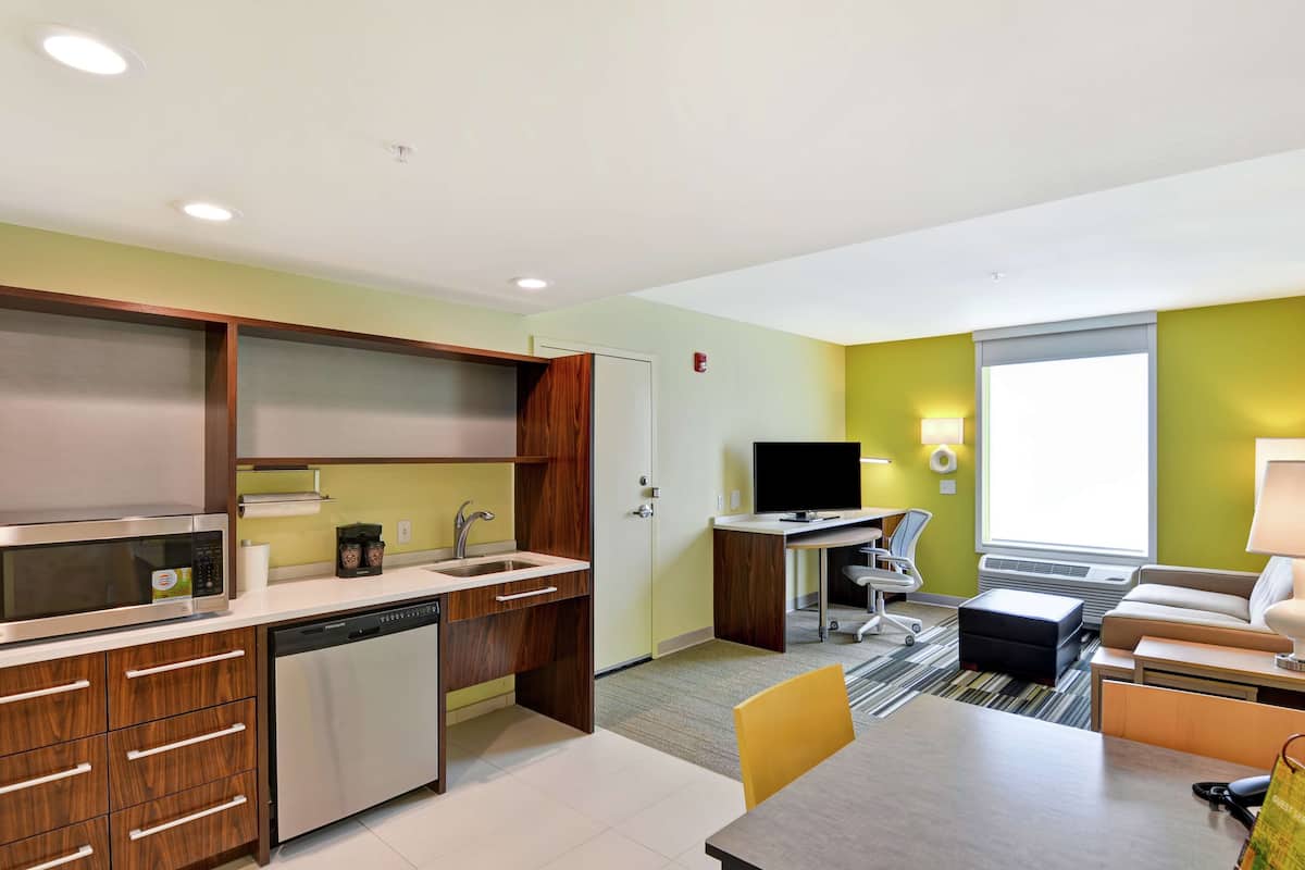 A compact hotel room with a kitchenette, a dining area, a TV, and a window with blinds.