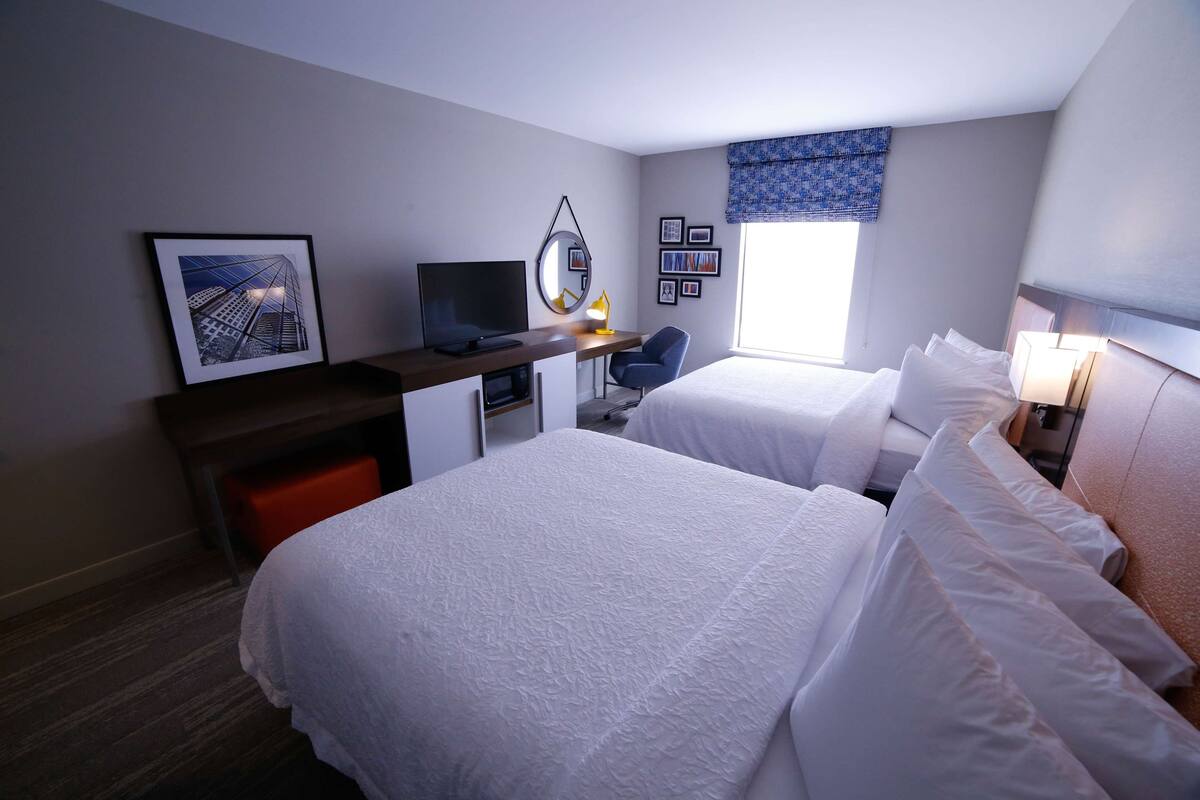 Ein Hotelzimmer mit zwei Betten, einem Schreibtisch mit Computer und Fernseher, einem kleinen Stuhl und einem Fenster mit Blick auf Gebäude.
