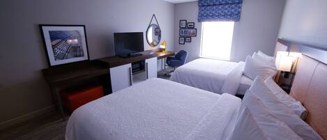 A hotel room with two beds, a desk with a computer and TV, a small chair, and a window with a view of buildings.