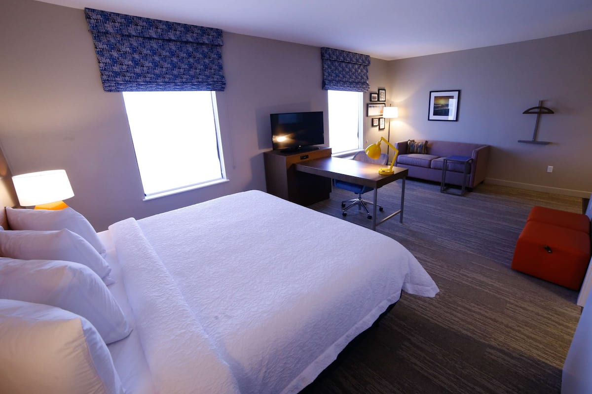 Ein Hotelzimmer mit einem großen Bett, einem Schreibtisch mit Computer, einem Fernseher, einer Couch und einem kleinen orangefarbenen Hocker.