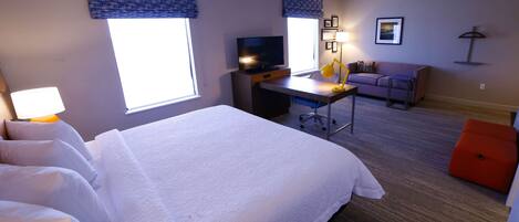 A hotel room with a large bed, a desk with a computer, a television, a sofa, and a small orange ottoman.