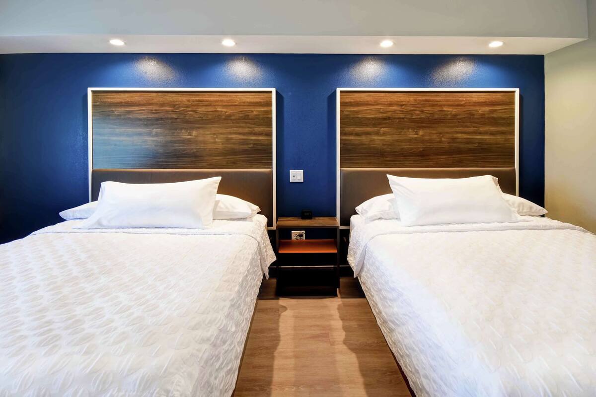 Habitación de hotel con dos camas, cada una con cabecero de madera, ropa de cama blanca y una mesita entre ellas.
