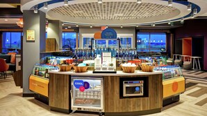 A hotel lobby with a central bar area featuring a variety of beverages and food items, a seating area with tables and chairs, and a large window providing a view of the outdoors.