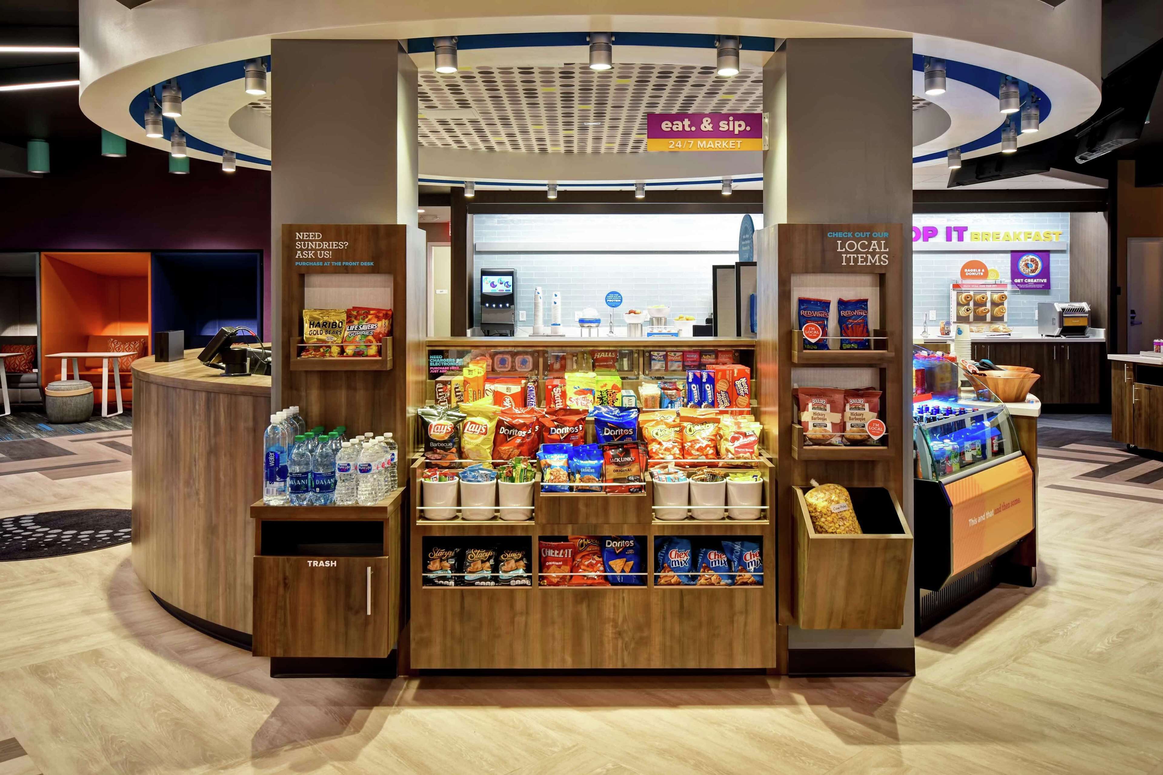 A snack kiosk with a variety of chips and bottled water, located in a modern indoor setting with a 24/7 market sign above.