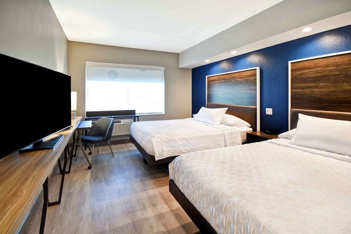 Habitación de hotel con dos camas, un escritorio, una silla, un televisor y una ventana con persianas.