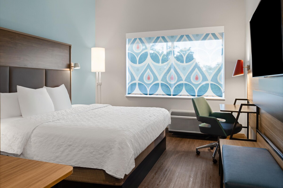 Una habitación de hotel moderna con una cama grande, un escritorio, una silla, un televisor y una ventana con un diseño geométrico.