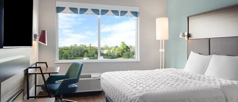 Habitación de hotel con cama, escritorio, silla y un ventanal con vista a árboles y casas.