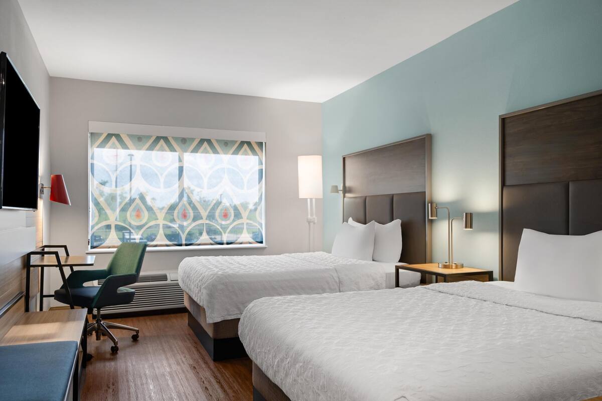 Ein Hotelzimmer mit zwei Betten, einem Schreibtisch, einem Stuhl, einem Fernseher und einem Fenster mit gemusterten Vorhängen.