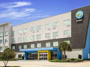 A modern hotel building with a blue and grey facade, a prominent 'tru by Hilton' sign, and a well-maintained entrance area with a palm tree and flower beds.