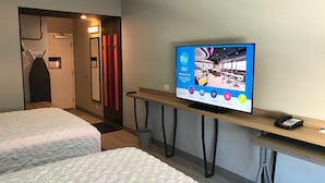 A hotel room with two beds, a TV displaying the hotel's amenities, a desk with a phone, and a door leading to the hallway.