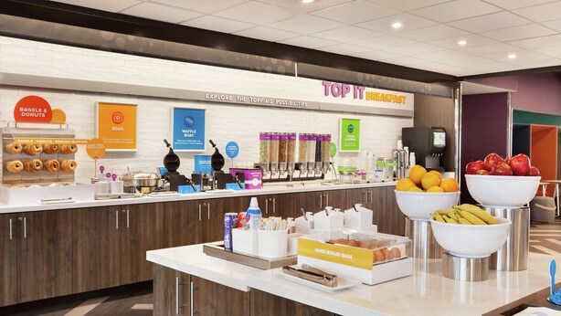 A breakfast buffet area with labeled food stations, including bagels, donuts, eggs, waffles, and a fruit bowl.
