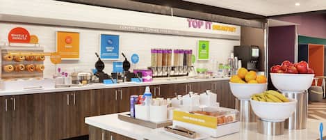 A breakfast buffet area with labeled food stations, including bagels, donuts, eggs, waffles, and a fruit bowl.
