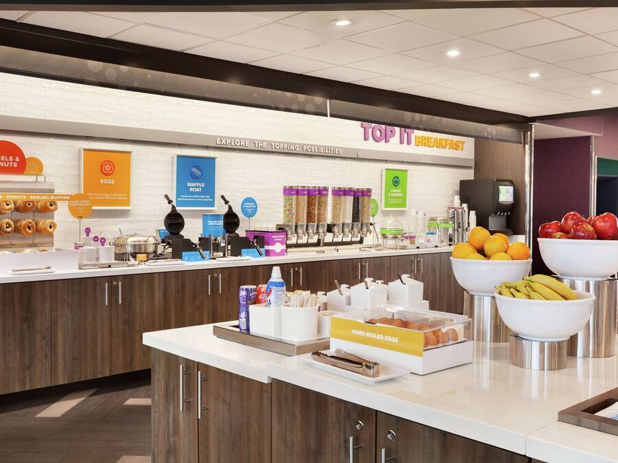 Zona de desayuno tipo buffet con estaciones de comida identificadas, que incluyen bagels, donuts, huevos, gofres y un cuenco de fruta.