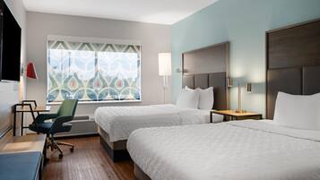 A hotel room with two beds, a desk, a chair, a TV, and a window with patterned curtains.