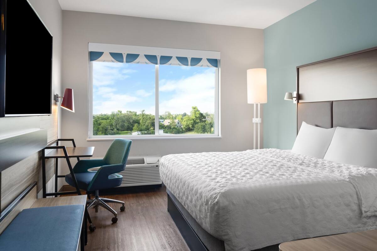 Ein Hotelzimmer mit Bett, Schreibtisch, Stuhl und einem großen Fenster mit Blick auf Bäume und Häuser.