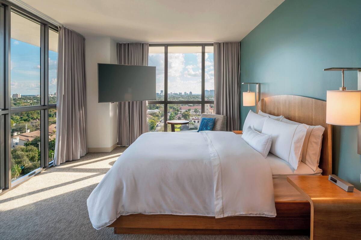 Une chambre d’hôtel avec un grand lit, un téléviseur fixé au mur, une table de chevet avec une lampe et une vue sur la ville grâce à de grandes fenêtres.