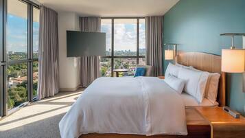 Habitación de hotel con una cama grande, un televisor montado en la pared, una mesita de noche con lámpara y vistas a la ciudad a través de amplios ventanales.