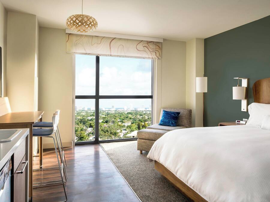 A modern hotel room with a large bed, a desk, and a view of the city through a large window.