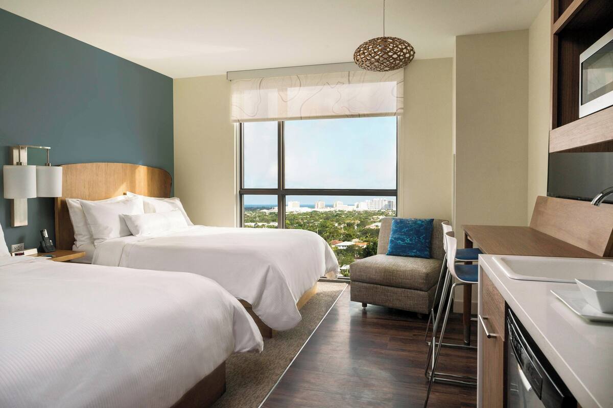 Une chambre d’hôtel avec deux lits, une kitchenette, un coin repas et une grande fenêtre donnant sur la ville.
