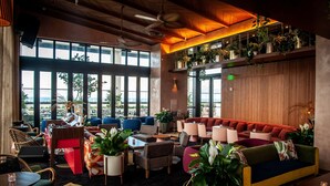 A modern lounge area with multiple seating options, including sofas and chairs, arranged around a central coffee table. There are large windows providing a view of the outdoors, and the space is decorated with various potted plants. - Element by Marriott Fort Lauderdale Downtown (Fort Lauderdale)