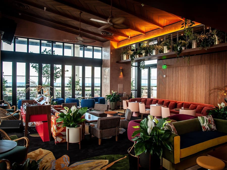 A modern lounge area with multiple seating options, including sofas and chairs, arranged around a central coffee table. There are large windows providing a view of the outdoors, and the space is decorated with various potted plants.