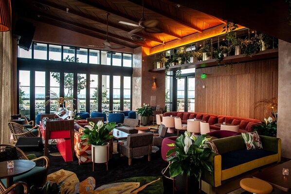 A modern lounge area with multiple seating options, including sofas and chairs, arranged around a central coffee table. There are large windows providing a view of the outdoors, and the space is decorated with various potted plants. - Element by Marriott Fort Lauderdale Downtown (Fort Lauderdale)