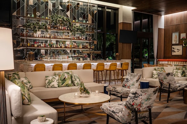 Un espace bar avec des étagères remplies de bouteilles et de verres, un coin salon avec des canapés et des fauteuils, et une grande fenêtre avec des rideaux.