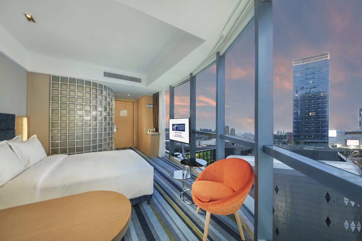 A hotel room with a bed, a desk, a chair, and a large window offering a city view.
