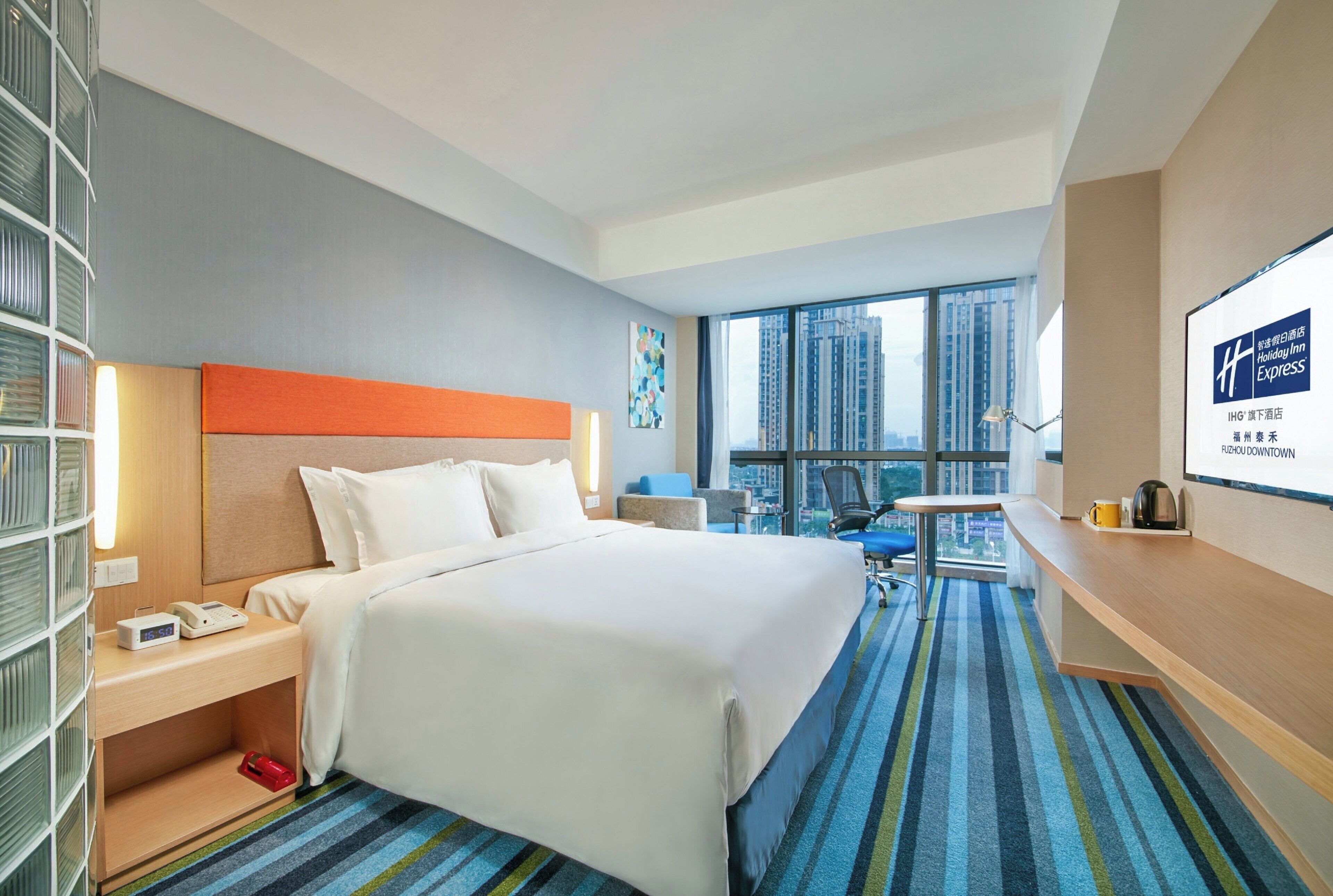 A hotel room with a bed, bedside table, lamp, and a view of the city.