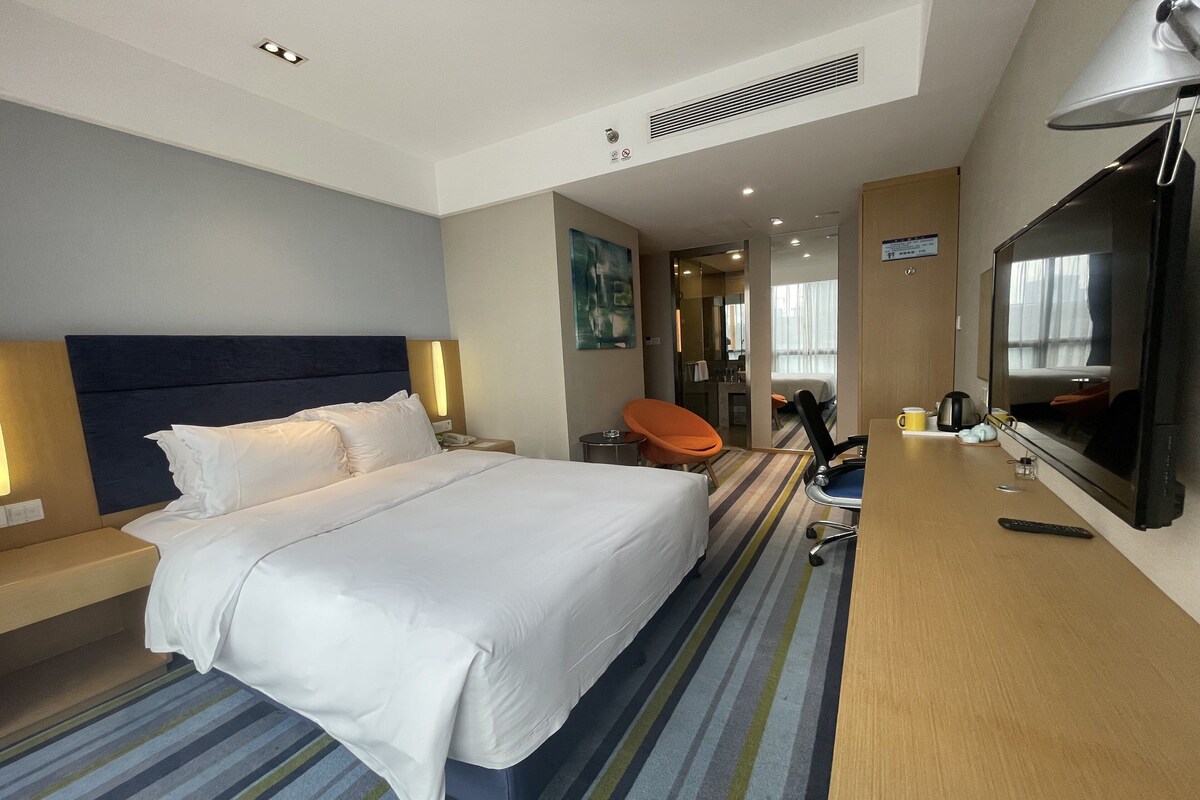 A hotel room with a large bed, a desk with a flat-screen TV, a chair, and a coffee maker.