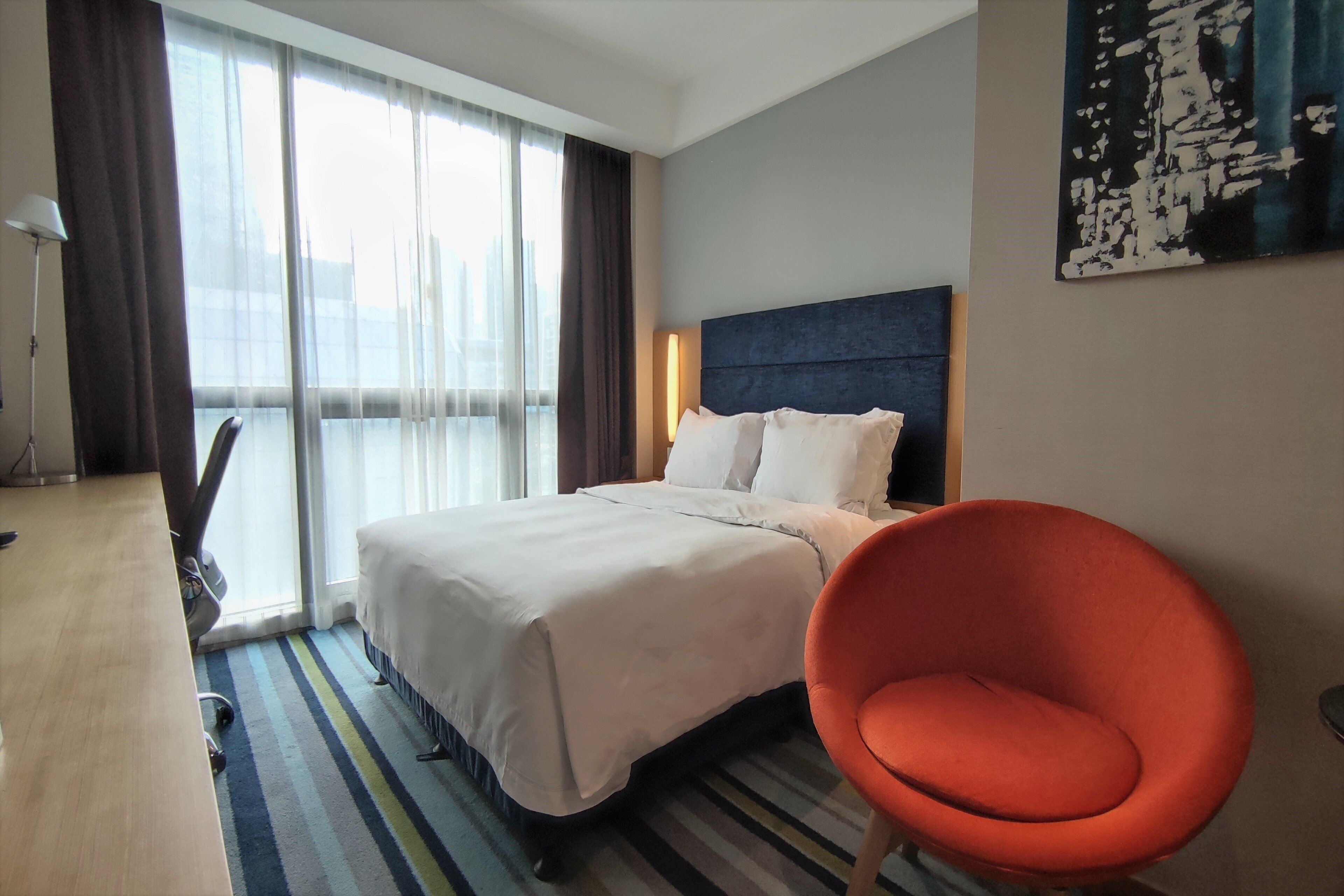 Una habitación de hotel con cama, escritorio, silla y un amplio ventanal con cortinas.