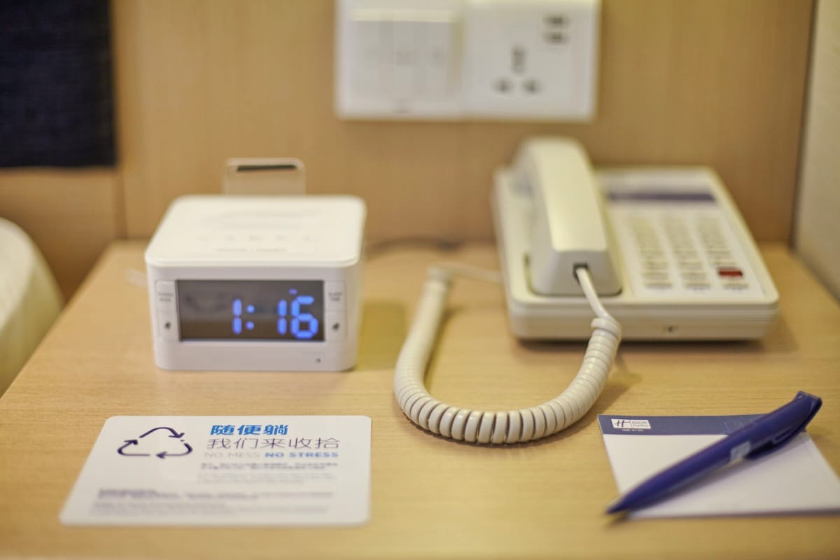 Une chambre d’hôtel équipée d’une horloge numérique affichant 1 h 16, d’un téléphone et d’une pancarte portant le symbole du recyclage et un texte.