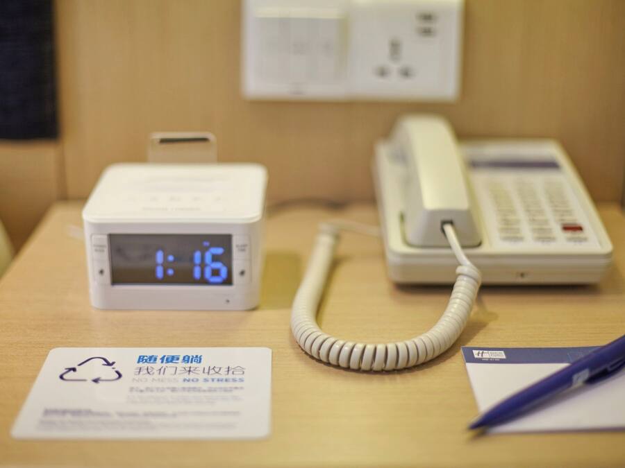 A hotel room with a digital clock displaying 1:16, a telephone, and a sign with a recycling symbol and text.