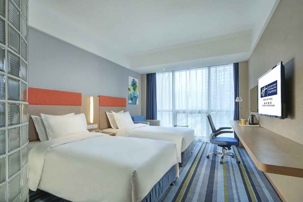 A hotel room with two beds, a desk, a chair, a TV, and a large window.