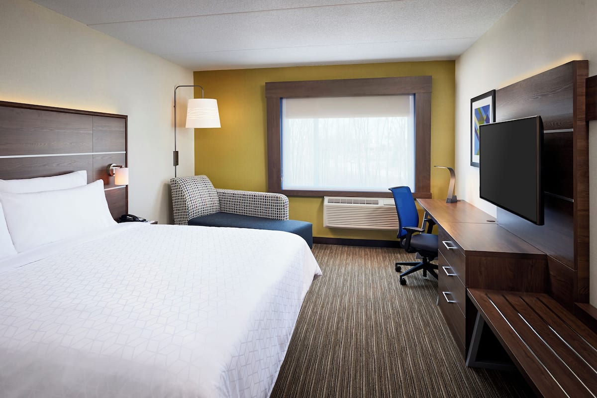 A hotel room with a large bed, a desk with a flat-screen TV, a chair, a sofa, and a window with blinds.