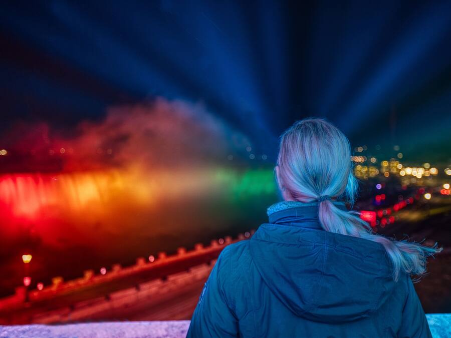 A person with a ponytail standing outdoors at night, facing a colorful light display.