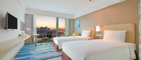 Ein Hotelzimmer mit zwei Betten, einem Schreibtisch, einem Sessel und einem Fernseher, mit Blick auf die Stadtlandschaft.