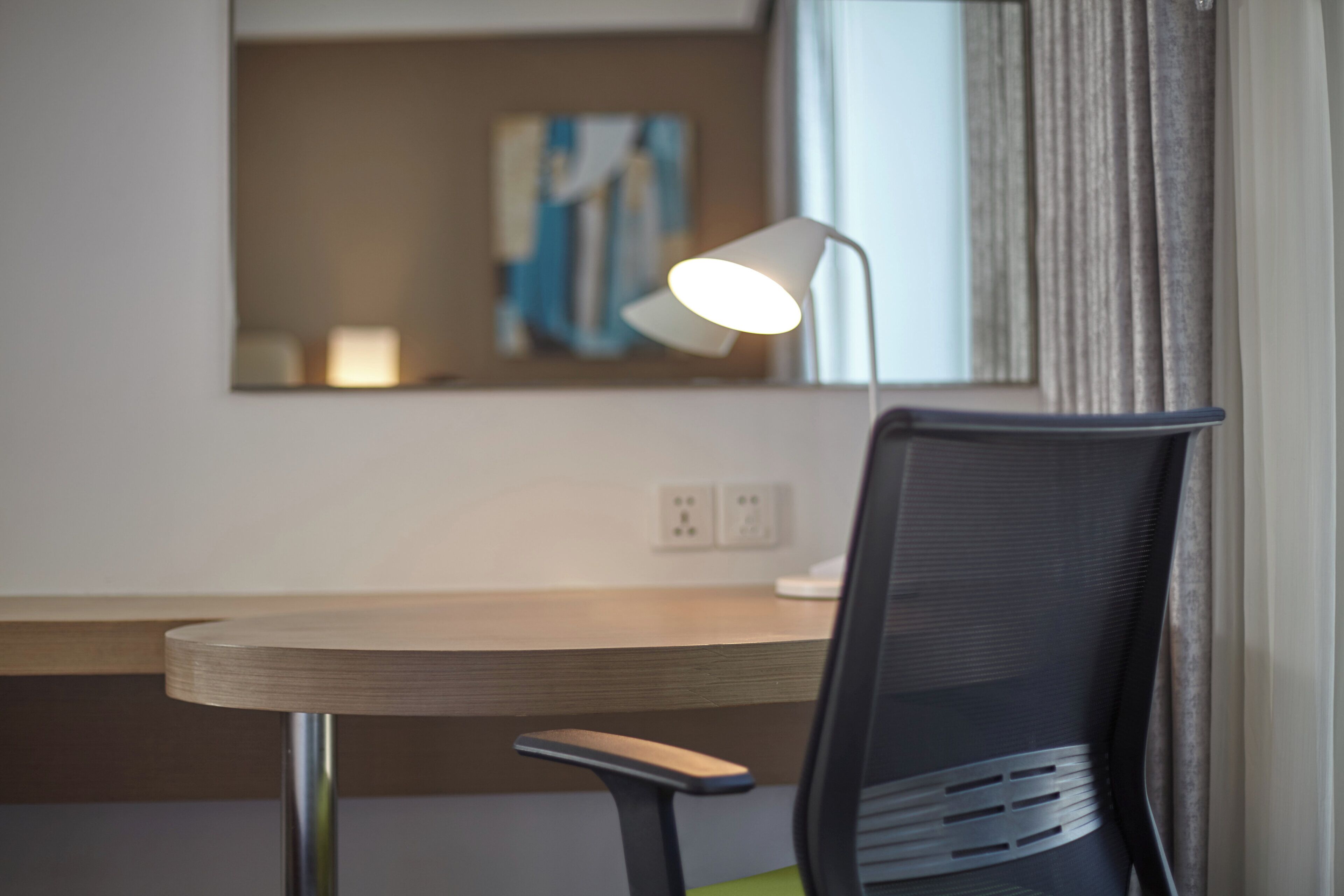 A hotel room with a desk, a chair, a lamp, a painting on the wall, and a window with curtains.