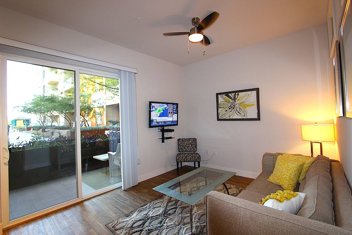 A living room with a glass table, a sofa, a television, and a ceiling fan.