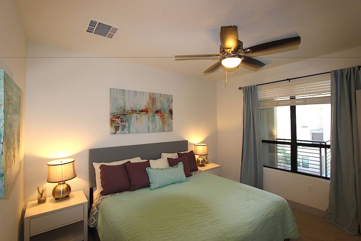 A bedroom with a bed, two bedside tables with lamps, a ceiling fan, a window with curtains, and a painting on the wall.