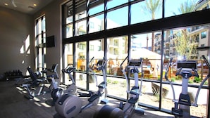 Un gimnasio con diversas máquinas de ejercicio, amplias ventanas y vistas a un edificio.