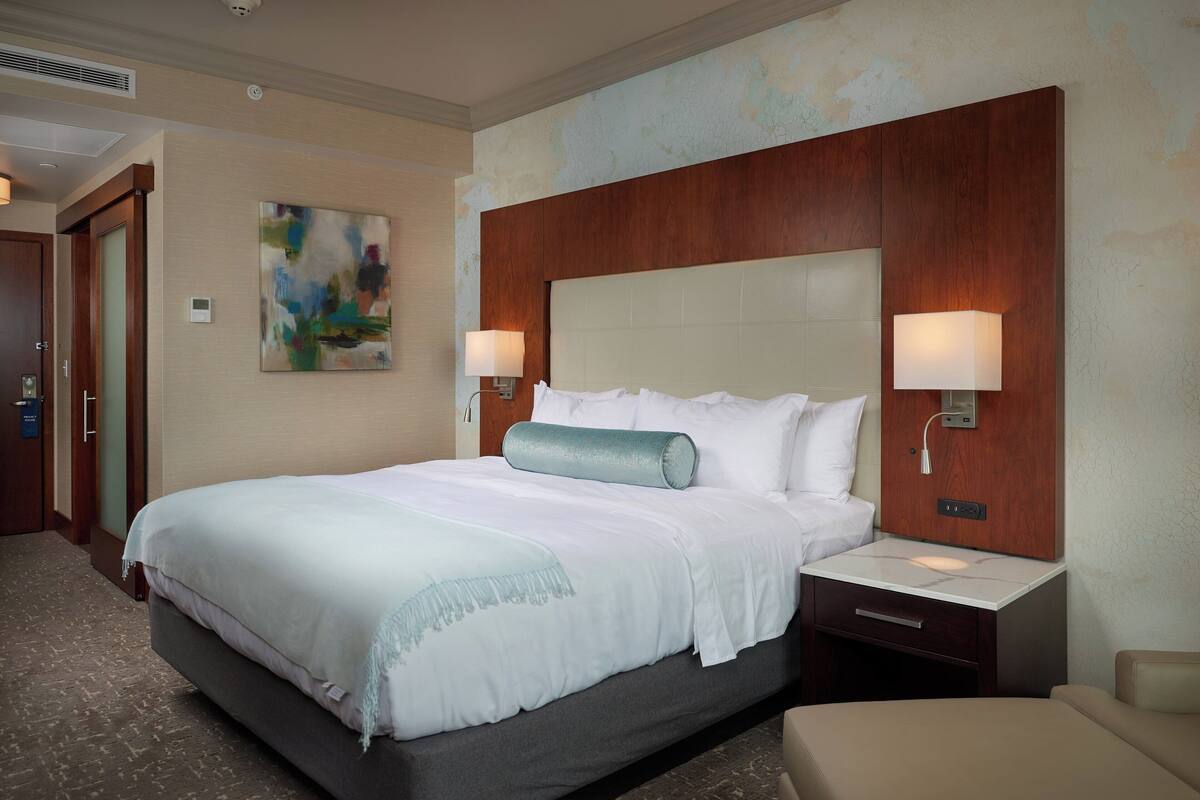A hotel room with a large bed, two bedside lamps, a wooden headboard, a painting on the wall, and a small nightstand.