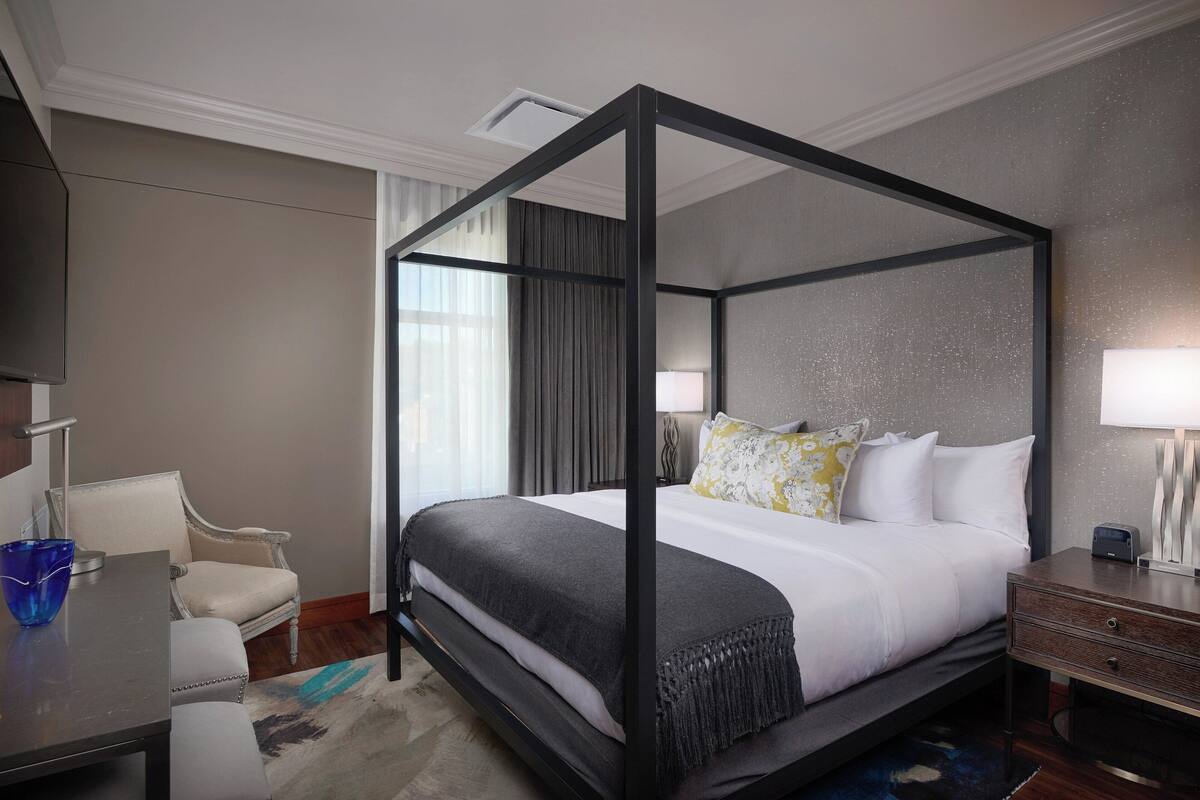 A four-poster bed with a grey bedspread and white pillows, a bedside table with a lamp, a chair, and a desk with a blue vase.