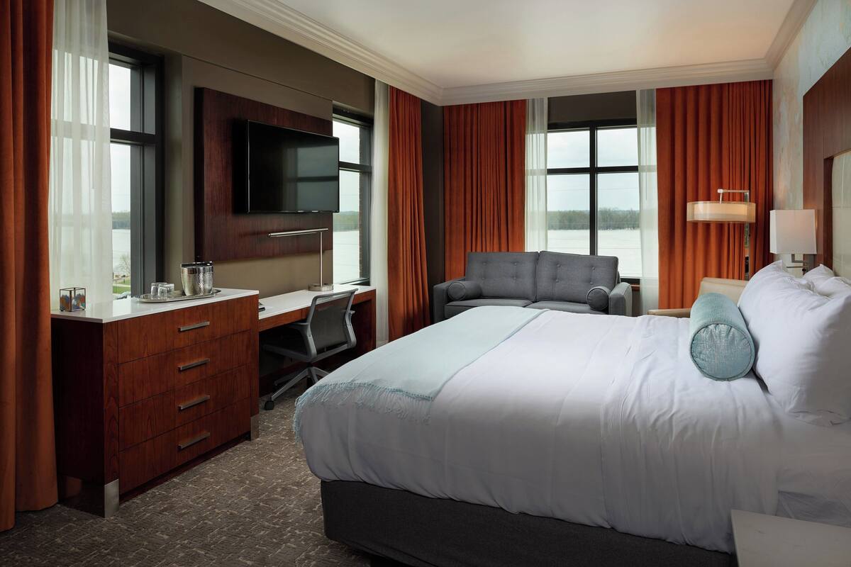 A hotel room with a large bed, a desk with a chair, a television mounted on the wall, and a window with curtains.