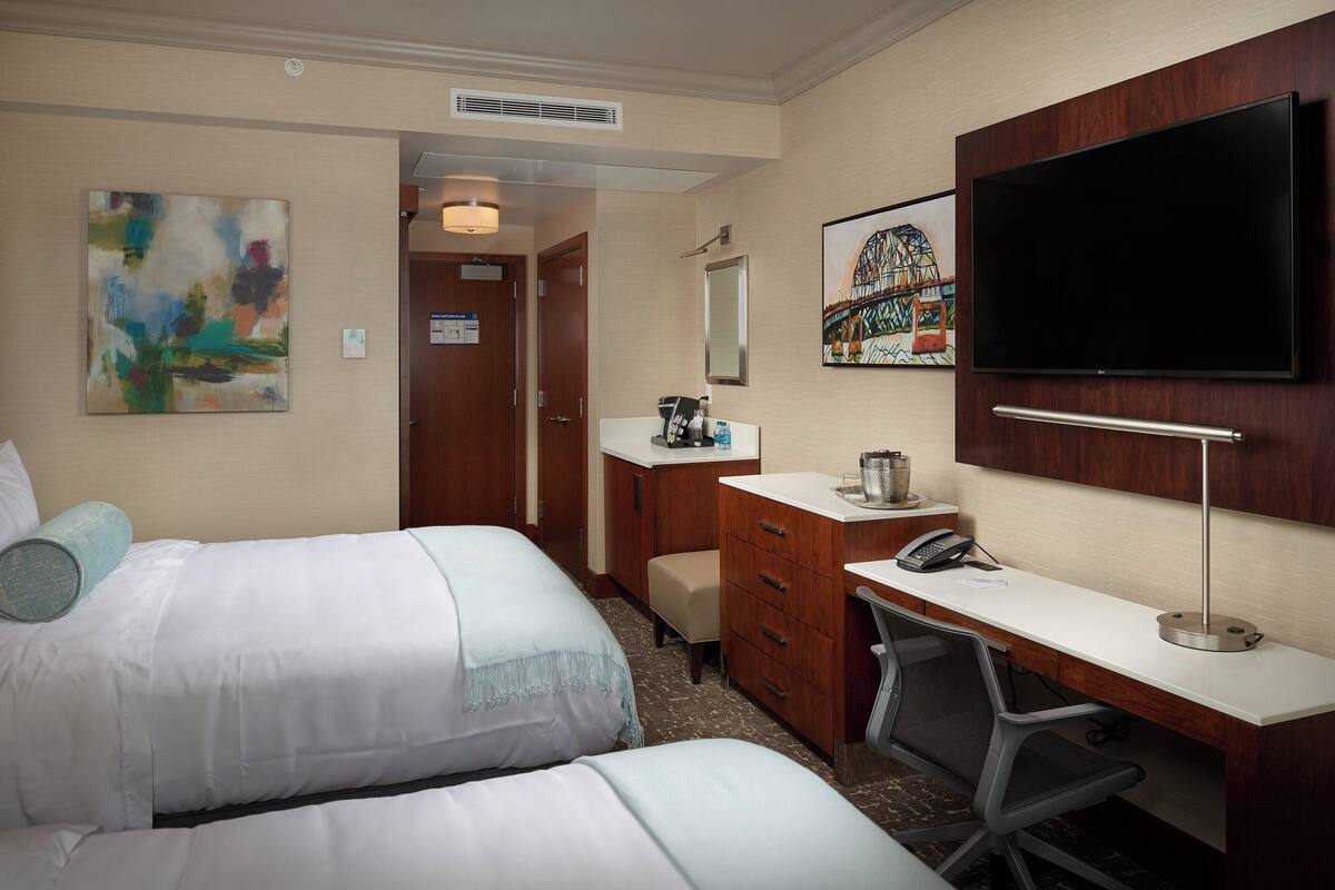 A hotel room with two beds, a desk, a chair, a TV, a desk lamp, a telephone, and a small cabinet.