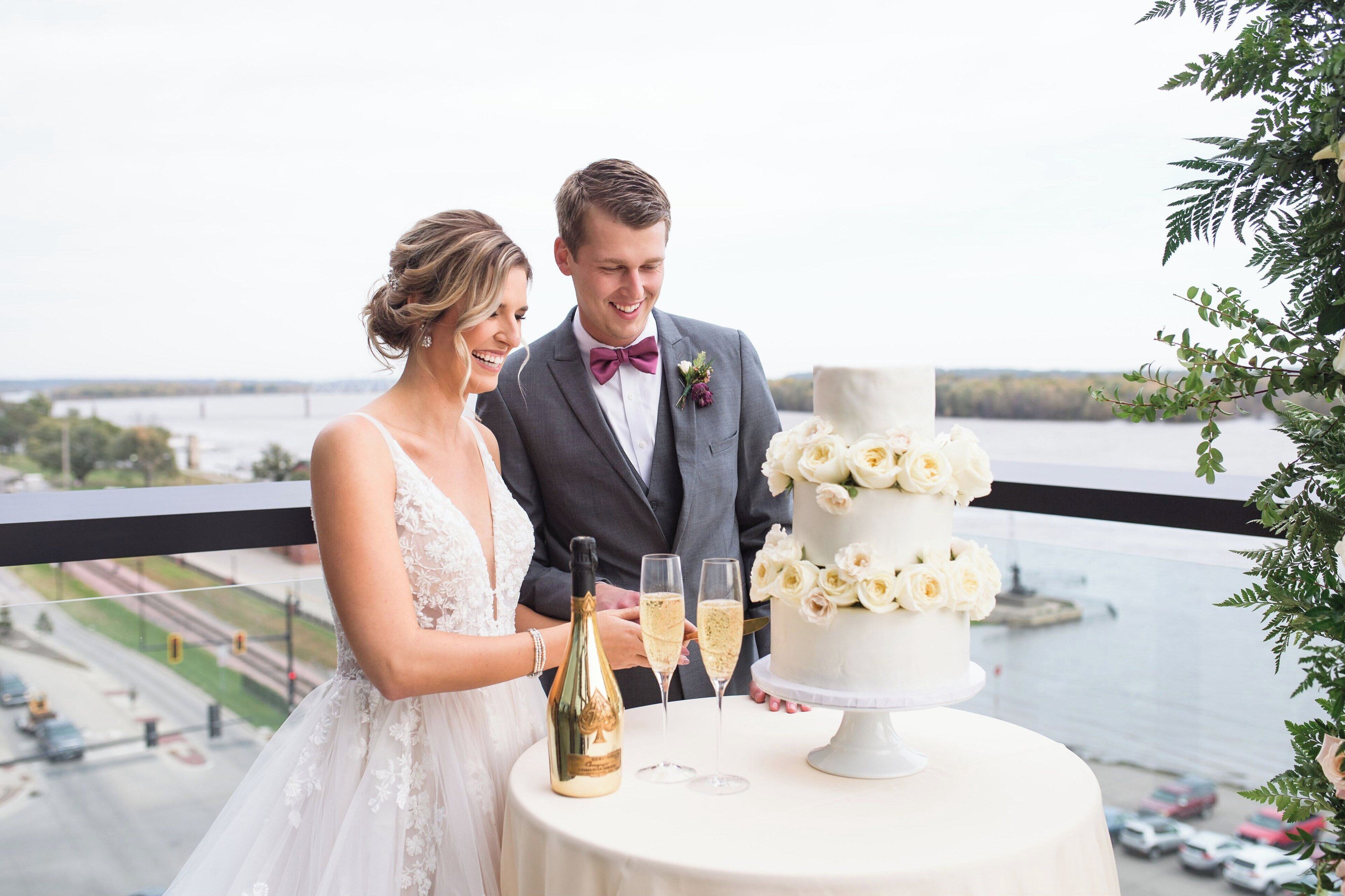 A couple cutting a white wedding cake with champagne on a balcony overlooking a body of water.