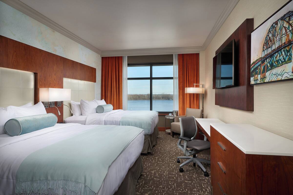 A hotel room with two beds, a desk, a chair, a TV, and a window with a view of the water.