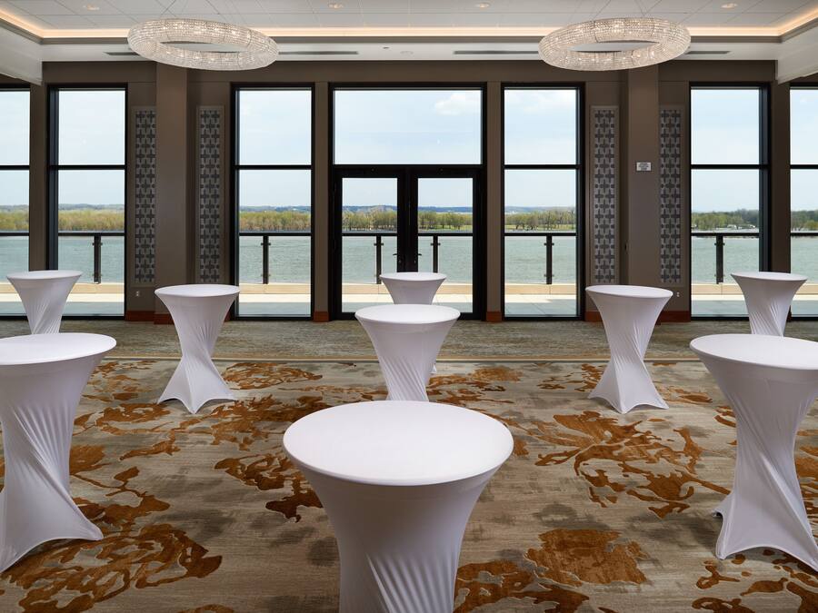 Een kamer met vijf ronde tafels met witte tafelkleden, grote ramen met uitzicht op een wateroppervlak, en een kamerbreed tapijt met dessin.